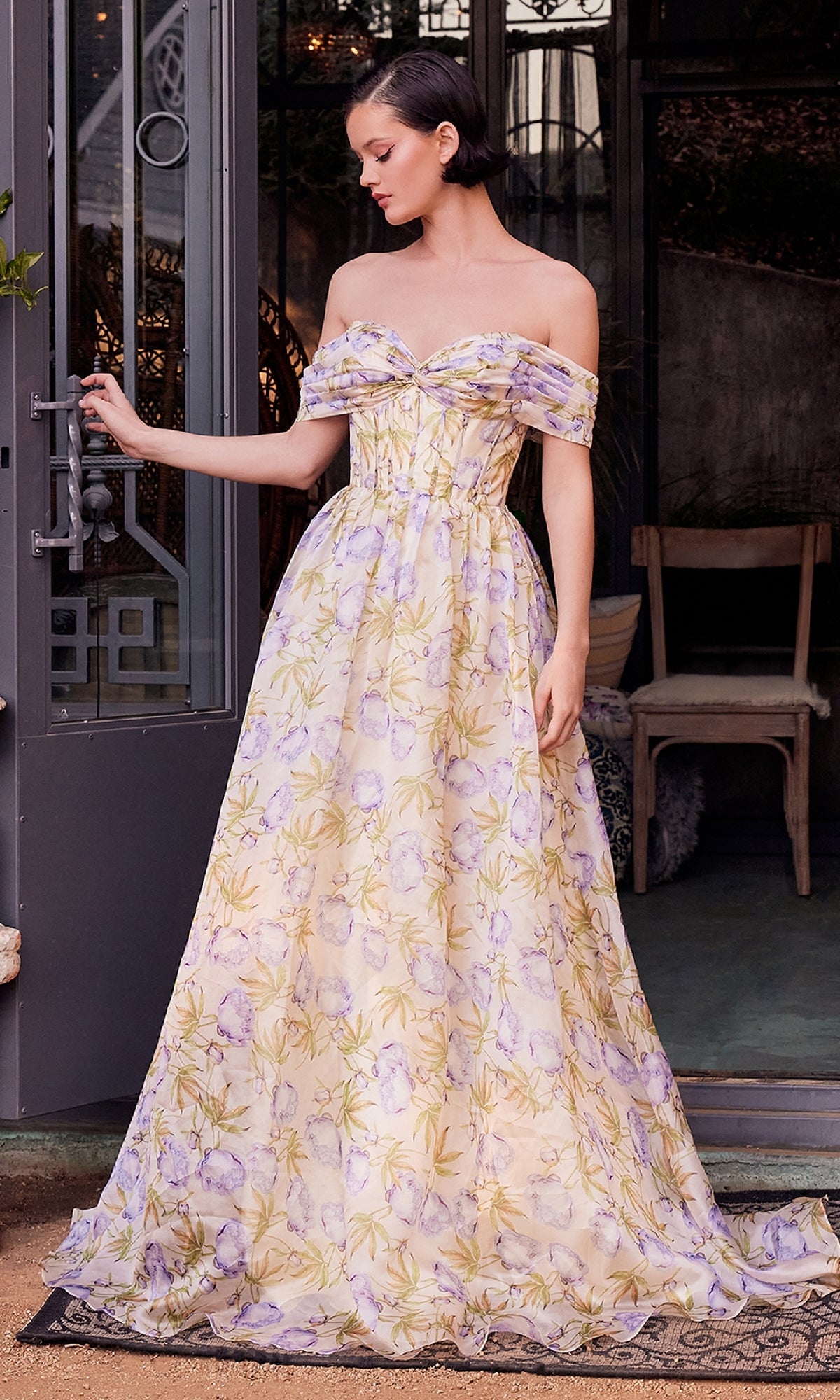 A woman poses in front of glass doors, wearing the Andrea & Leo A1466—a floor-length, off-the-shoulder floral-print prom dress with a soft corset-style bodice.